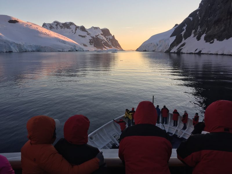 A ship navigating through the Lemaire Channel at sunset with people watching from the bow